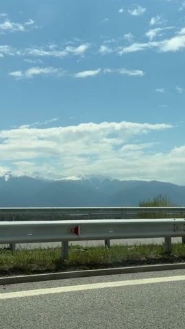 The video shows a view from a moving vehicle, likely a car, traveling on a highway. A silver guardrail is visible along the right side of the road. Beyond the guardrail, there's grassy vegetation and a view of distant mountains. The mountains are partially covered in snow. The sky above is blue with scattered white clouds. The road itself is asphalt with painted white lines.