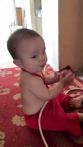 A baby is sitting on a red rug, wearing red pants and a red ribbon around its neck. The baby is clapping its hands, and it appears to be holding something small and dark in its right hand. The baby's expression is one of concentration and slight amusement. The background is blurry but appears to be an indoor setting with a wall and some furniture. A white cord is visible on the rug near the baby.