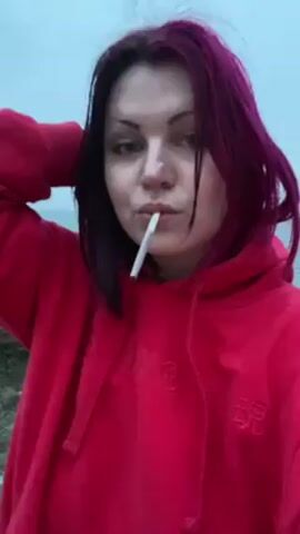 A woman with dyed magenta hair and a red hoodie is seen in a close-up shot. She has a cigarette in her mouth and is holding a lighter, presumably to light it. The background shows a body of water, possibly the sea, with a cloudy sky. She adjusts her hair with her left hand. Her right hand is tattooed.