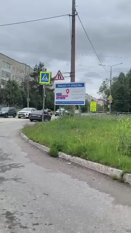 The video captures a street scene on a cloudy day, likely in a residential area. The camera pans across a road with several cars driving and stopped at a traffic light. On the left side of the road are apartment buildings and trees. On the right side of the road, there is a pedestrian crossing with a traffic signal, road signs, and more trees. Further down the road, a large mural is visible on the side of an apartment building, depicting a colorful, abstract design and a face. The ground appears wet in places, suggesting recent rain. There are no clear interactions between people visible in the footage.