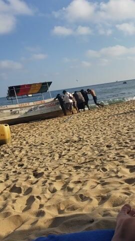 This image depicts a group of men launching a boat from a sandy beach into the ocean, likely in Mazunte, Mexico. Approximately seven men are actively pushing the long, narrow vessel. Most are positioned on the sandy shore, exerting force on the boat's side, while one man is already in the shallow water, pushing from the front.

The boat, painted white with blue accents, clearly advertises its services with text including "SPORT FISHING," "TOURS SNORKELL," "CORAL," and "SURFING," indicating its role in local tourism and recreation. A flag with horizontal red, yellow, and green stripes is visible on the boat's front. The beach consists of light-colored sand, bearing indentations from the boat's movement. A yellow object is partially visible in the bottom right corner.

The scene unfolds under a bright, clear blue sky with scattered white clouds, suggesting a sunny day, likely morning or early afternoon. The ocean is calm with small waves breaking near the shore, and several seagulls are seen flying in the distance over the water. The activity appears to be a routine preparation for a day of sea excursions, characteristic of coastal life and tourist offerings in locations like Mazunte.