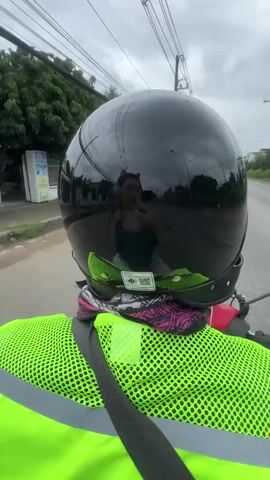 The video shows a first-person perspective of someone riding a motorcycle. The rider is wearing a black helmet, a neon green safety vest, and a pink and black bandana around their neck. The helmet's reflective visor shows a reflection of the rider, who is a woman with cat-ear headphones and appears to be taking a selfie with her phone. The surroundings are a paved road with trees and utility poles visible. The sky is overcast.