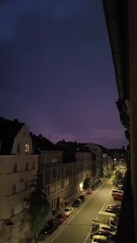 This is a vertical, low-angle, nighttime exterior shot looking down a street lined with buildings and parked cars. The sky is a deep purple, transitioning to a lighter purple near the horizon. The street is illuminated by streetlights, casting a warm glow on the asphalt and the parked vehicles. The buildings on either side of the street are mostly multi-story structures with visible windows, some of which are lit from within. Several cars are parked along the street, facing in various directions. The perspective suggests the shot is taken from a window or balcony overlooking the street. There are no people visible in the shot, and no significant actions or interactions are occurring. The overall atmosphere is quiet and urban.