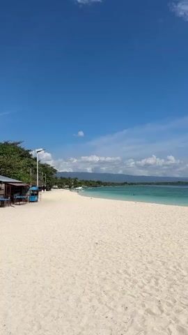 A person is visible in the water on the right side of the frame, appearing to swim or float. In the distance, the coastline is lined with trees and vegetation, with mountains visible further inland. The sky is a clear blue with scattered white clouds. The water transitions from a light turquoise near the shore to a deeper blue further out. The beach is covered in fine, white sand, with a few dark objects, possibly towels or bags, scattered on the sand near the waterline. On the left side of the frame, there is a small building with a brown roof and some blue structures in front of it, possibly seating or stalls. A lamppost is also visible on the left.