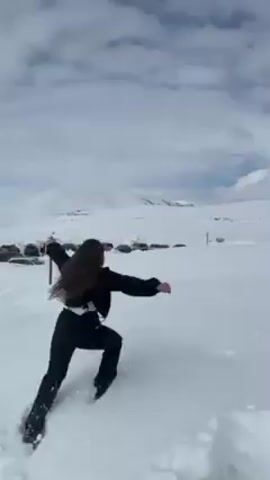 A woman with long dark hair, wearing a black jacket and pants, walks through deep snow, arms outstretched for balance. The background features a snow-covered landscape with mountains in the distance under a cloudy sky. Footprints mark her path in the snow.