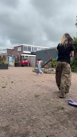 Two people are outdoors on a paved area. A child is running away from the camera while a woman, facing away from the camera, appears to be holding a bat or a stick. They are near a building and a car.