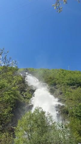 The video shows a multi-tiered waterfall cascading down a steep, rocky hillside covered in lush green trees and vegetation. The water appears white and frothy as it plunges down the rocks. The sky above is a clear, bright blue. Power lines are visible in the upper portion of the frame. There are also branches and leaves in the upper right and lower left corners of the frame. The camera remains static, capturing the scene from a distance.