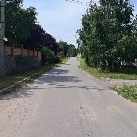 The video shows a paved road in a rural or suburban area. The road is lined with trees and fences on both sides. The fence on the left is made of stone and brick, while the fence on the right appears to be made of wood or concrete. The sky is partly cloudy. The camera appears to be moving forward, suggesting the video is a recording of someone driving or walking down the road. There are no people or animals visible in the video. The overall atmosphere is peaceful and quiet.