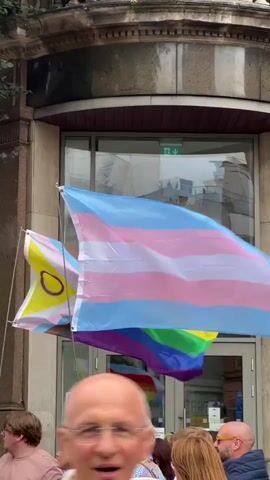The video shows two flags waving in front of a building with large glass doors. The flag on the right is a transgender pride flag, featuring horizontal stripes of light blue, pink, and white. The flag on the left is yellow with a purple circle in the center, representing the intersex flag. Below these flags, a rainbow flag can be seen through the glass doors. Several people are visible in the foreground, their faces partially obscured, suggesting they are part of a crowd or event. The building's facade is made of stone and has decorative architectural elements. The lighting suggests an outdoor daytime setting.