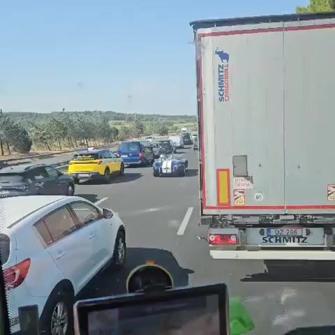 A blue convertible sports car with white racing stripes is driving on a highway in moderate traffic. The driver is wearing a white helmet and a white shirt. Other vehicles visible include a large white semi-truck on the right, a yellow taxi on the left, a blue minivan, and various other cars and trucks. The highway appears to be in a rural or semi-rural area with trees and hills in the background under a clear blue sky.