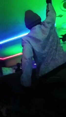 A video shows a person in a black dress dancing in a room lit with colorful neon lights. The room has red, green, and purple lighting along the ceiling. Initially, another person in a patterned shirt and hat is shown. The dancing person raises their arms and moves to the music. Other people are partially visible sitting in the background.