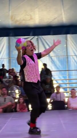 A short, dark-haired person in a pink sequined vest and black pants, red socks, and large black shoes juggles a multi-colored ball while standing on a purple stage. The juggler tosses the ball into the air, catches it, and throws it behind their back. They are performing in front of an audience sitting in red chairs. The background is a blue and white tent.