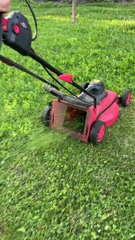 A person's hands are shown operating a red and black electric lawnmower across a green lawn. The lawnmower is actively cutting the grass, with clippings visible being ejected from the side. The camera angle is low and close to the ground, emphasizing the action of the lawnmower. The environment appears to be a residential yard, with grass covering the majority of the frame.