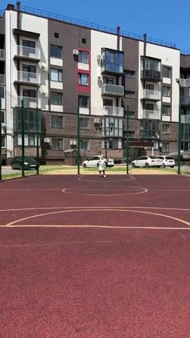 A young child runs toward the camera on a red outdoor basketball court. The child is wearing a light green dress and white shoes. In the background, a multi-story building with a brown and white facade stands under a clear blue sky. Several cars are parked in front of the building. The court has yellow lines marking its boundaries and a net in the center.