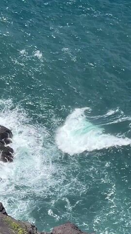 The video shows a rocky coastline from a high vantage point. Waves crash against the dark rocks below, creating white foam against the turquoise water. The camera then pans out to reveal a long stretch of coastline with steep, rocky cliffs dropping into the ocean. The sky is blue with a few scattered clouds. The overall scene is one of natural beauty and ruggedness. The wind is blowing, as suggested by the camera movement.