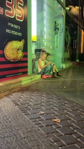 Two young girls are sitting on a curb outside a shop. The girl on the left is wearing a white tank top, blue jeans, and pink sandals. She has her hair pulled up in a bun and is looking at a phone. The girl on the right is wearing a leopard print shirt and brown boots. She also has her hair pulled up and is looking at a phone. They are both focused on their phones and appear to be interacting with each other. The shop behind them has a glass front and is illuminated with green lights. The ground is paved with cobblestones, and there are a few fallen leaves on the ground. A poster advertising pizza is visible on the left side of the shop. The scene is dimly lit, suggesting it is nighttime or early evening.