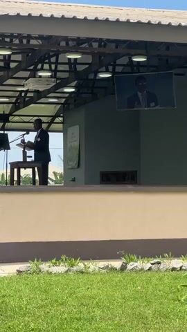 A man in a suit and tie stands behind a wooden podium on a raised stage. He is gesturing with his left hand and holding a book or papers in his right. There is a screen displaying a person in a suit and tie above and to the right of the stage. The stage is covered by a roof with exposed beams and lights. In front of the stage is a beige wall and beyond that, a grassy area with rocks and some plants. The background shows trees and buildings.