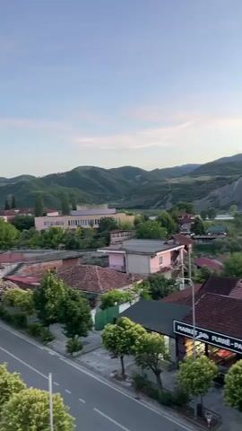 The video is a vertical, handheld shot looking out over a small town or village at dusk. The sky is a soft blue with streaks of orange and yellow from the setting sun. Rolling green hills, dotted with trees, form the backdrop. In the foreground, there are several buildings with red tile roofs and some newer, modern structures, including a white apartment building. A road runs along the bottom of the frame, and trees line the sidewalk. A small market or shop with text above it is visible on the right side of the frame. The scene is quiet and peaceful.