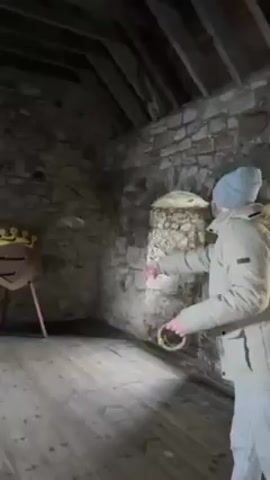 A person in a beige jacket and grey knit cap throws a set of rings towards a shield target in a stone room. The target is brown and features a gold crown on top. The shield is set on three legs, and the person throws rings at it but misses each time. The room is mostly stone with wooden floors and a wood beam ceiling. There is an archway in the wall behind the target.