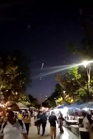 The video captures a nighttime scene of a bustling outdoor market, likely a festival or celebration, where fireworks are being displayed overhead.  The perspective is from within the crowd, looking up towards the sky where a bright red firework explodes. The market is lined with stalls, possibly selling food or crafts, and illuminated by strings of lights and overhead streetlights. People of varying ages stroll along the walkway, enjoying the festivities. The trees on either side of the path add a natural element to the urban setting. The sky is a deep blue, characteristic of twilight or early evening.