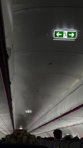 The video shows the interior of an airplane cabin from the perspective of a passenger. The camera is pointed upwards towards the ceiling, where emergency exit signs are visible. The aisle is visible with rows of seats on either side, occupied by passengers. There appears to be some smoke or mist emanating from the ceiling in the middle of the cabin. The overall lighting is dim, consistent with the cabin lights being on during a flight.