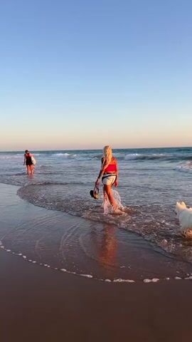 This video shows two people walking in the ocean with a white dog. The sky is clear and blue, and the sun is setting, casting a warm glow over the scene. The water is shallow and clear, and small waves are lapping at the shore. The first person, a woman with blonde hair, is wearing a red top and white shorts and is walking towards the ocean. She is holding a pair of sandals in her hand. A white dog is walking alongside her. The second person, a woman with dark hair, is further out in the ocean and is wearing a dark dress. She is also walking towards the ocean. The beach is sandy and appears to be unpopulated.