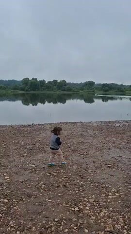 A child stands on a pebble beach facing a large body of water. The water is calm and reflects the overcast sky and a tree-lined shore in the distance. The child is wearing a grey jacket and light-colored pants. The child turns slightly to their left and then begins to run away from the water towards the right of the frame. The child's feet are visible as they run. There are no other people or animals visible in the immediate vicinity of the child. The environment appears to be a natural landscape, possibly a lake or riverbank.