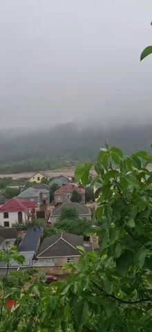 A panoramic drone shot captures a village nestled in a valley on a foggy day. The village is characterized by numerous houses with colorful roofs, varying in architectural styles and shades of red, gray, and blue. Dense green foliage surrounds the village, with trees and shrubs peeking through the buildings. In the background, a mist-shrouded mountain range rises, its slopes covered in trees. The atmosphere is subdued due to the heavy fog, lending a serene yet slightly melancholic feel to the scene. The camera slowly pans across the landscape, revealing more of the village and the surrounding natural beauty. There are no people or visible animals in the frame, the focus being entirely on the tranquil village and its misty environment.