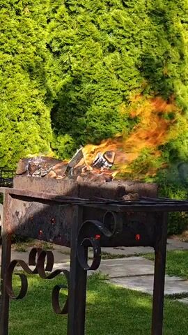 A time-lapse video shows a barbecue grill with a fire burning in it. The grill is made of metal and has a decorative design on the legs. In the background, there is a lush green hedge. The fire burns intensely, with flames reaching upwards and flickering. The video captures the process of the fire growing and consuming the wood in the grill.