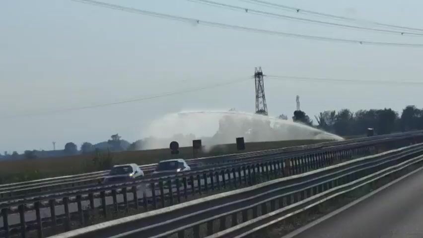 A wide-angle view of a field with a large sprinkler system spraying water is visible across a highway. The sprinkler is in the middle ground, with its water spray reaching high into the air and fanning out over a large area of the field. In the background, power lines and utility poles are visible. The highway is in the foreground, with several cars driving on it. The sky is clear and bright, suggesting it is daytime. The overall scene depicts an agricultural landscape with a focus on the irrigation system.