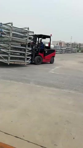 The video shows a red and black forklift moving a large stack of silver-colored metal frames in an outdoor, industrial setting. A person is operating the forklift. The area is paved with concrete and has other stacks of metal frames in the background. The sky is overcast.