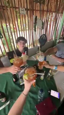 A group of four people are seated around a table in an outdoor setting, possibly a restaurant, and are holding up their drinks in a toast. The table is covered with a green tablecloth, and there are drinks, bottles, and a phone on the table. The drinks appear to be alcoholic beverages, garnished with limes. One person is smiling at the camera. Behind the group is a bamboo or thatched wall, with a "no smoking" sign visible. A small fan is also present. The overall impression is one of a casual, social gathering.
