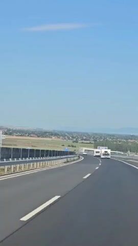 The video shows a view from a vehicle traveling on a highway. The highway has two lanes in the direction of travel, separated by a solid white line. The right lane has a white dashed line to its right, while the left lane has a barrier. The highway is bordered by a metal guardrail on the right and a concrete barrier on the left. In the distance, there are signs on the right side of the highway, and a few buildings and trees can be seen. Two white vehicles, possibly campers or RVs, are traveling in the same direction ahead of the camera. The sky is clear and blue, with a single wispy cloud. The overall environment is a sunny day on a highway, with agricultural or suburban landscape in the background.