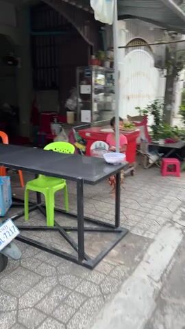 A young child wearing a pink shirt and patterned pants stands on a paved outdoor area. The child is looking to the right of the frame. In front of the child is a black table with a bowl of food on it. To the right of the child is a red cooler on wheels. In the background, there is a building with an open doorway and a glass display case. The scene appears to be a street or sidewalk outside a food stall or restaurant.
