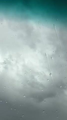 The video shows a view through a car windshield on a rainy day. Large raindrops are visible on the glass, obscuring the grey, cloudy sky behind them. The top of the windshield has a green tint. Ambient music is playing in the background.