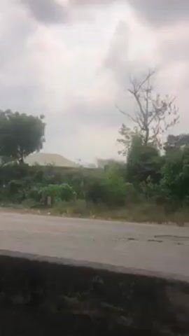 The video shows a shot of a grey asphalt road and some greenery on a cloudy day. The shot seems to be taken from behind a low concrete wall. On the other side of the road, there is a hillside covered with trees and bushes. A couple of buildings are visible in the background, including a light-colored one with a light-colored roof. A car appears briefly on the road. The sky is overcast with heavy grey clouds.