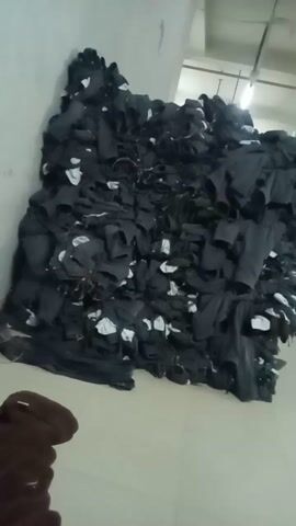 Here's a forensic description of the video:

**Scene:** An indoor industrial setting, likely a garment factory or warehouse.

**Objects:** A large, chaotic pile of dark gray or black clothing items is the primary focus. The clothes are stacked haphazardly, reaching almost to the ceiling. The items appear to be individual pieces, possibly shirts or pants. Small white tags or labels are visible within the stack.

**Environment:** The floor is a light gray or beige color.  The walls are a pale, off-white. There is a metal pipe or railing system attached to the ceiling. In the upper part of the frame, a green industrial structure or machine is partially visible.

**Action:** There is no immediate action. The video appears to be a static shot of the pile of clothes.