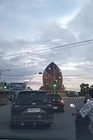 The video shows a street scene in Sigli, Indonesia, as traffic moves around a large, distinctive roundabout monument. The monument, shaped like a large seed or pod with a wooden exterior, dominates the background. It has a unique, curved form and appears to be a significant landmark.

Several vehicles are visible on the road, including black cars and motorcycles. The perspective is from inside a vehicle, looking forward. A motorcycle with two people, a driver and a passenger wearing helmets, is in the foreground, moving away from the camera. The passenger wears a light-colored head covering and a backpack.

Traffic lights are visible, with at least two showing green, indicating traffic is flowing. The sky is overcast with dramatic clouds, suggesting either dawn or dusk. The overall lighting is somewhat dim, consistent with these times of day.

On the left side of the road, a billboard or large sign is partially visible, featuring images of what appear to be cars. In the background, beyond the roundabout, more buildings and trees can be seen, typical of an urban or suburban environment. The word "SIGLI" is visible on the roundabout monument, confirming the location. The mood is neutral, depicting a routine moment of urban transit under a cloudy sky.