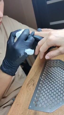 The video shows a person, presumably a manicurist, wearing black gloves and a light-colored uniform, applying a clear liquid from a small white bottle to the fingernail of another person. The other person's hand is also covered in a black glove. A cotton pad is held in the gloved hand of the manicurist. A metal grate rests on a wooden table in front of the hands.
