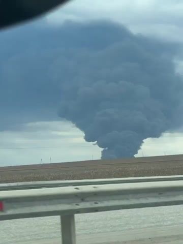 The video shows a large plume of black smoke rising in the distance, as viewed from a car traveling on a highway. A dark blue dump truck passes in the foreground on the left, partially obscuring the view initially. The highway has a metal guardrail with red reflectors.  A field lies between the highway and the smoke. Another car, a dark-colored SUV, passes in the opposite direction.  Power lines and a road sign are visible near the smoke. The sky is overcast.
