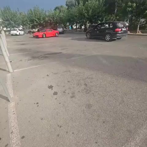 A black SUV is parked on the right side of a paved lot. A red sports car is driving in the lot. Several trees line the background of the lot. The sky is cloudy.