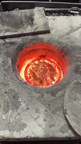 The video captures a close-up, top-down view of a circular furnace opening in an industrial setting. The furnace is constructed from a rough, gray, possibly refractory, material, showing signs of wear with some cracks and darker stains on its surface.

The central focus is the intensely glowing interior of the furnace, which radiates a bright, fiery orange-red light. Within this circular opening, a crucible or melting pot is visible, filled with what appears to be molten metal or metal pieces in the process of melting. The material inside the pot is a vibrant, sparkling orange and yellow, displaying a turbulent and shimmering surface, indicating extremely high temperatures. The inner walls of the furnace surrounding the pot also glow with a deep red hue.

A metallic strip or bar is partially visible on the upper left edge of the furnace's outer structure. No individuals are present in the frame, and there is no discernible text. The audible background noise is a loud, consistent humming or whooshing sound, characteristic of an operational high-temperature industrial furnace or a powerful burner. The overall scene depicts an active metal melting or refining process within a workshop or foundry environment. The location context provided is Saint-Rémy-lès-Chevreuse, France.