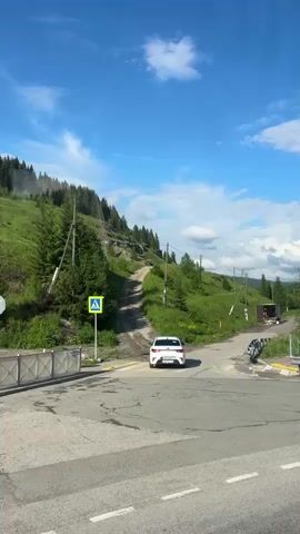 A white car is turning at a crossroads on a paved road. A pedestrian crossing sign is visible on the left side of the road. The road leads to a dirt road that ascends a grassy hill covered in trees. A white truck with its back doors open is parked on the dirt road, with some items visible in the back. The sky is blue with scattered clouds. The overall environment appears to be a rural or mountainous area.