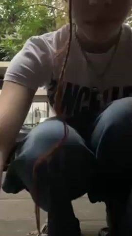 A young woman wearing a white t-shirt, baggy jeans, and sandals is standing on a sidewalk in an urban environment. Two cars are parked on the side of the road. The woman does a short dance move, then a man walks in front of her wearing a brown skirt and no shirt, covered in tattoos. The woman puts her hands on his shoulders, as he is now crouched in a squat.