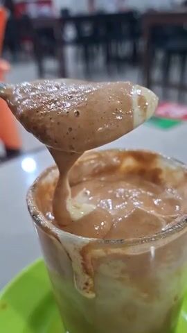 A close-up shot captures a spoon filled with a frothy, light brown beverage, possibly coffee or a milkshake, suspended over a clear glass of the same drink. The beverage appears thick and creamy, with some liquid dripping down the side of the glass, suggesting it is cold or contains ice. The glass is resting on a bright green saucer. The background is blurred, showing hints of a dining establishment with tables and chairs, indicating the drink is likely in a cafe or restaurant. The lighting is artificial and appears to be from overhead sources.