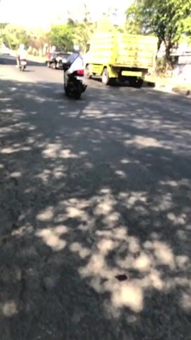 The video captures a street scene with several vehicles and pedestrians. In the foreground, there is a paved road with shadows of trees cast across it. Towards the middle ground, a yellow truck is driving away from the camera. To the left, several motorcycles are also visible, some carrying passengers, and a white car is parked on the right side of the road. The environment appears to be a sunny day with clear skies and trees lining the street, suggesting an outdoor, possibly urban or suburban, setting.