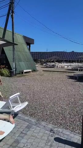 The video shows a view of a gravel courtyard in front of a white building with a dark roof. The building has several windows and a door with the number 8 above it. Several white lounge chairs are arranged in front of the building. In the background, a large field of solar panels is visible under a clear blue sky. A few umbrellas are scattered among the solar panels. The camera is panning to the right.