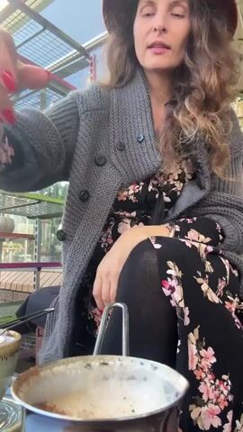 A woman wearing a hat and a grey cardigan over a floral dress sits outdoors at a table with a coffee cup and a metal sieve. She holds the sieve over the cup and appears to be filtering something into it. The background shows a greenhouse-like structure with plants and shelves.