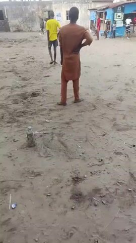 The video shows a sandy area with people standing around in the background. A man in a brown tunic and pants stands in the foreground with his hand on his hip. There is a dirty plastic bottle on the ground to his left. Other people are walking around in the background, and there are some buildings in the distance.