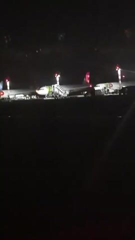 The video shows three airplanes parked on an airport tarmac at night. The airplanes are illuminated by lights from the ground and from the airport's navigation systems. Stairs are extended to the doors of two of the planes, suggesting passengers may be boarding or disembarking. The background is dark, with some distant lights visible, possibly from other airport structures or the city. The overall scene depicts a typical nighttime operation at an airport.